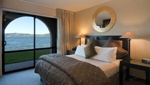 Una habitación de hotel con una cama grande, dos mesitas de noche con lámparas, un espejo redondo y vistas al agua a través de un ventanal amplio.