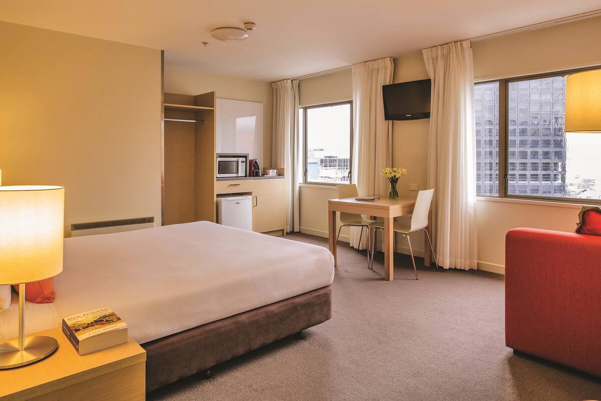 Ein Hotelzimmer mit einem Bett, einem Schreibtisch, einem Sessel, einem Fernseher und einem Fenster mit Blick auf die Stadt.