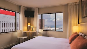 Une chambre d’hôtel avec un grand lit, un bureau avec une lampe, une chaise et une vue sur le paysage urbain grâce à de grandes fenêtres.