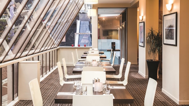 Un espace de restauration moderne dans un hôtel, avec une longue table dressée pour quatre personnes, un escalier doté d’une rampe en verre et une grande fenêtre offrant une vue sur la ville.