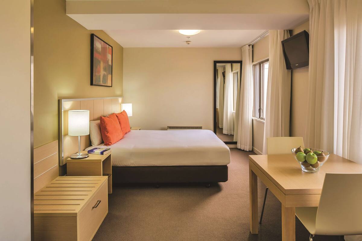Ein Hotelzimmer mit Bett, Nachttisch mit Lampe, Schreibtisch mit Obstschale und einem an der Wand befestigten Fernseher.