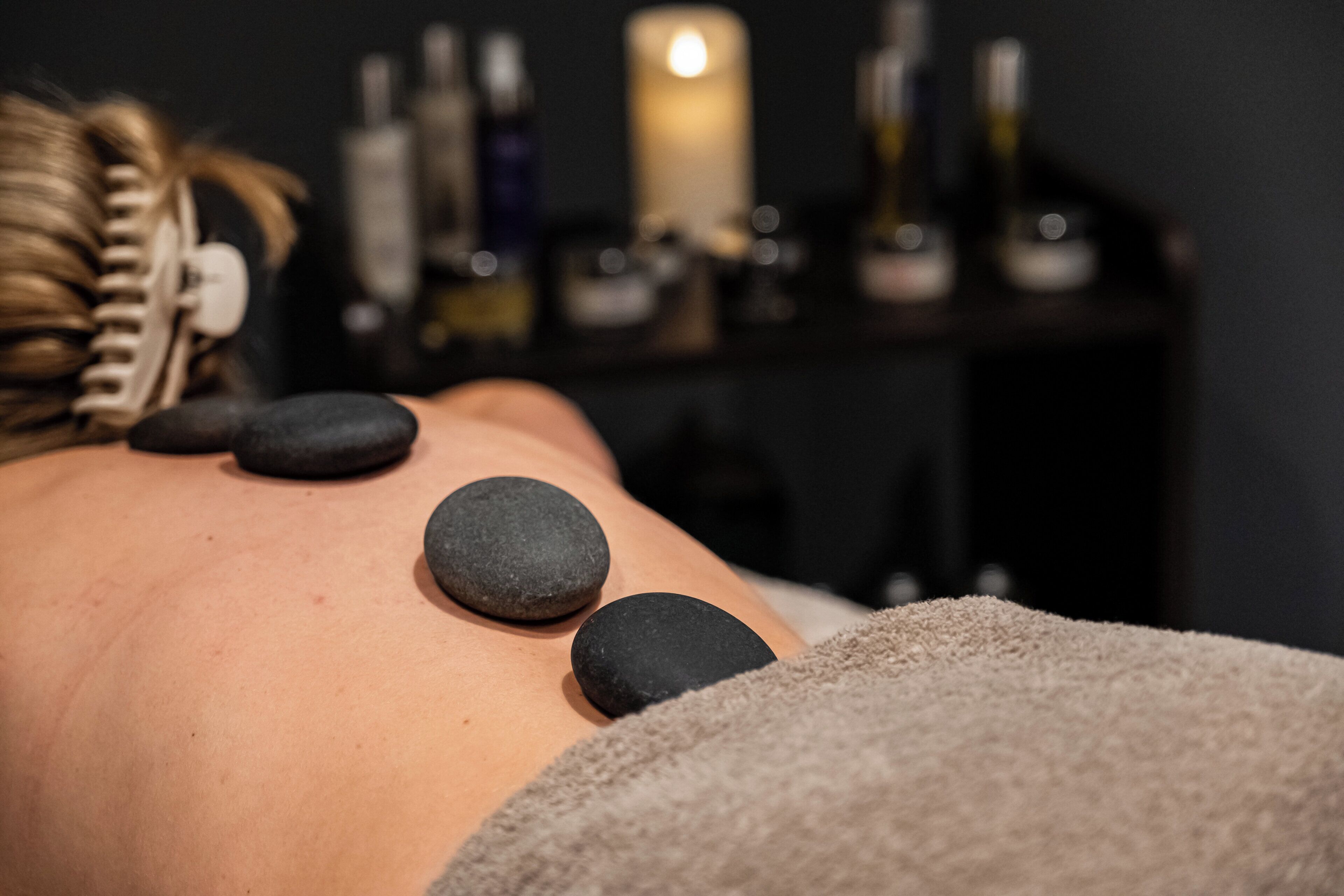 Iemand ontvangt een steenmassage, met drie stenen op de rug, een kaars en verschillende flessen op de achtergrond.