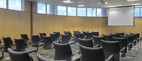 A conference room with rows of black chairs, a large projection screen, and windows with a view of the sky.