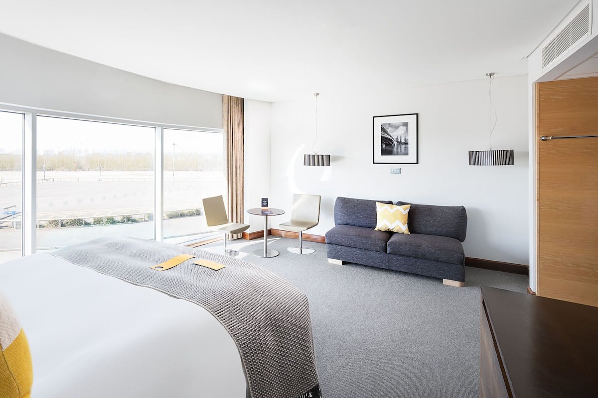 A modern hotel room with a large bed, a grey sofa, a small round table, and a view of a parking area through large windows.