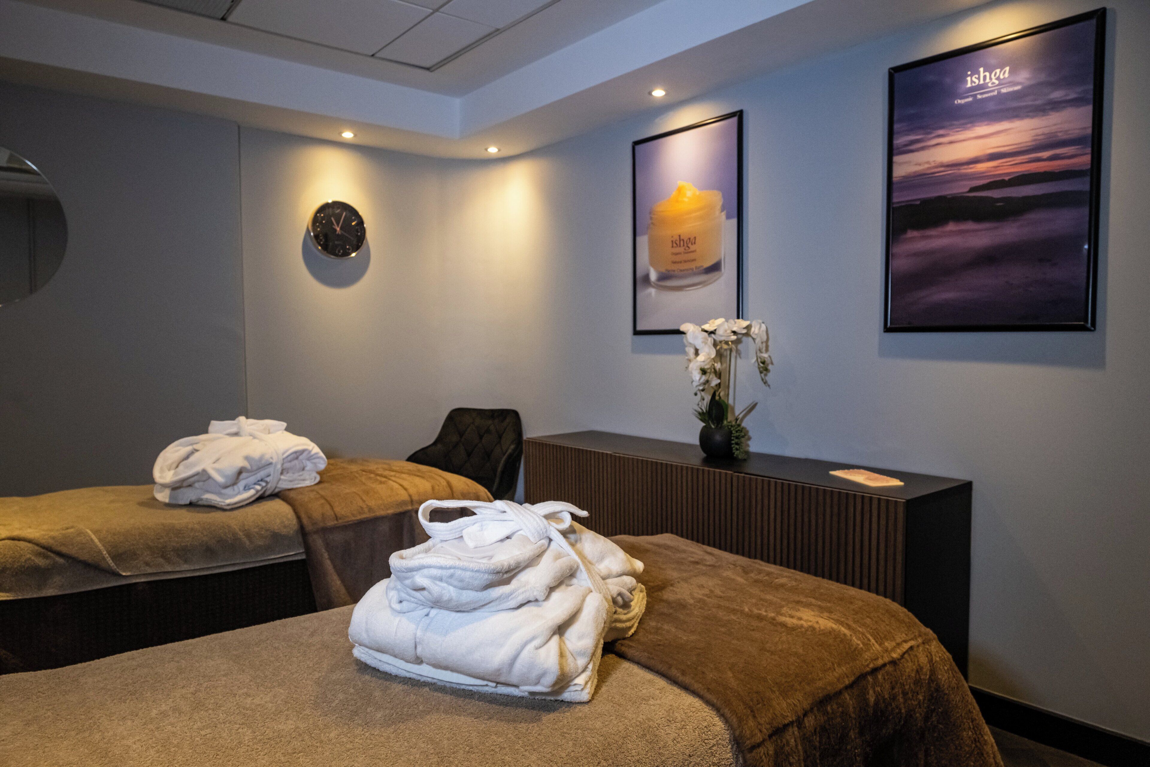 A spa room with two massage tables, towels, a clock, and framed artwork on the wall.