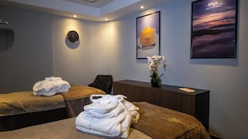 A spa room with two massage tables, towels, a clock, and framed artwork on the wall.
