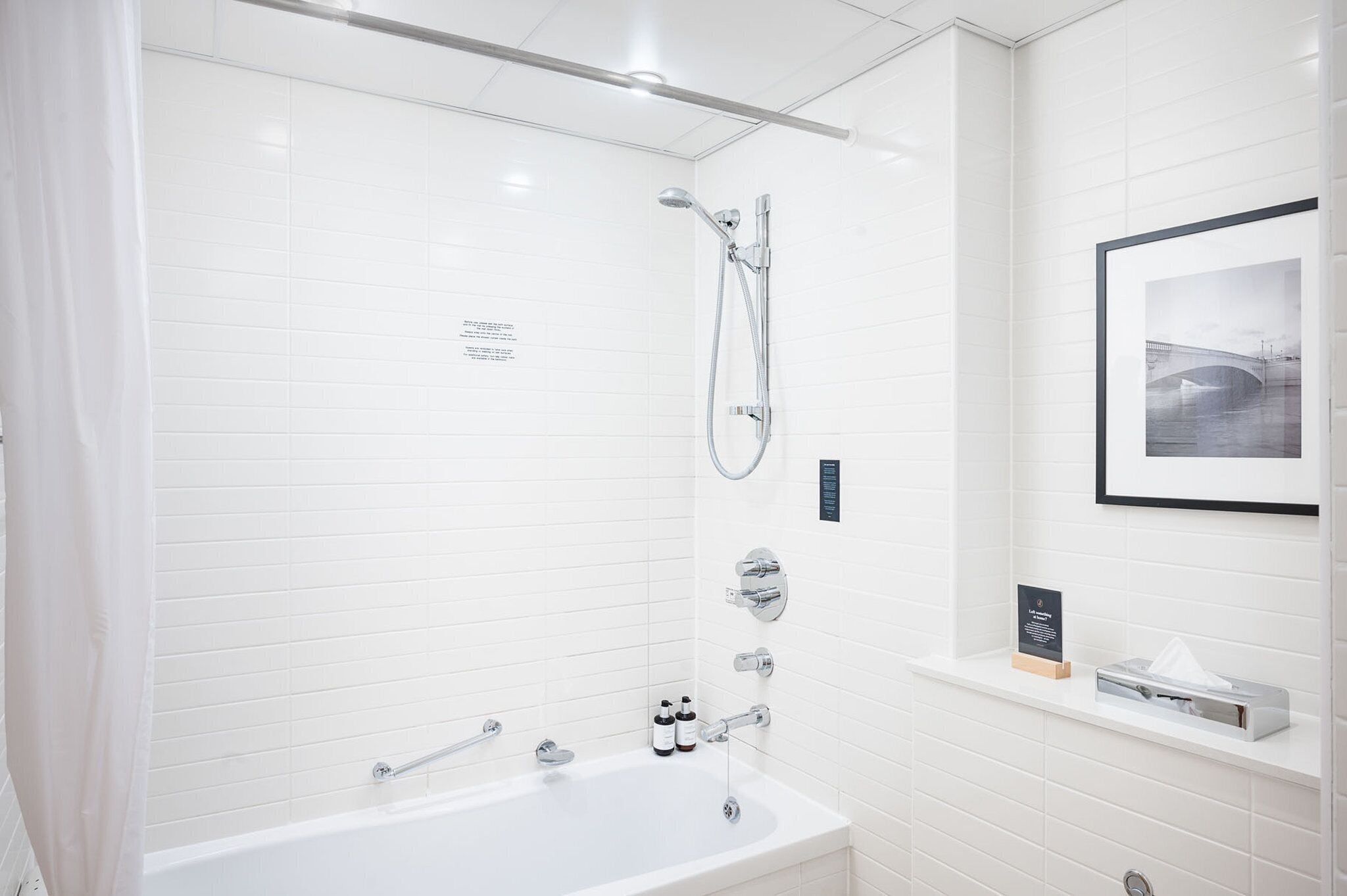 A modern bathroom with white tiled walls, a bathtub, a shower with a handheld showerhead, and a framed picture on the wall.