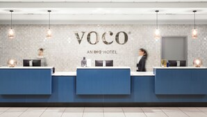A hotel reception area with a modern design, featuring a reception desk with a digital screen, a logo above the desk, and a tiled wall.