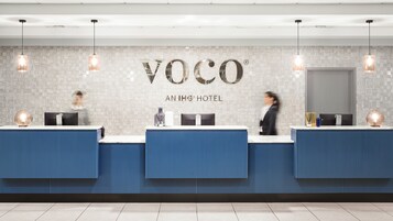 A hotel reception area with a modern design, featuring a reception desk with a digital screen, a logo above the desk, and a tiled wall.