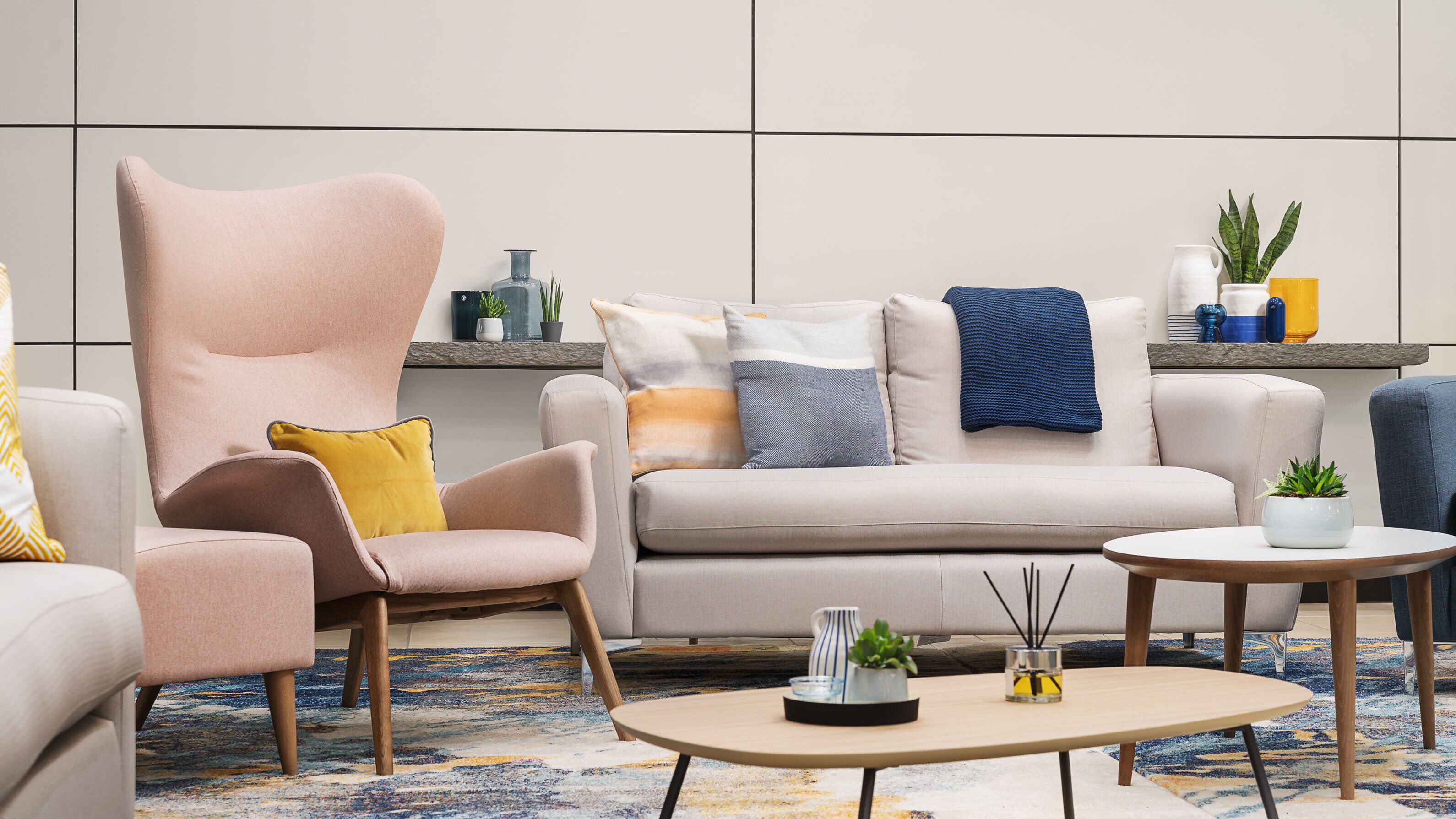 A modern living room with a large armchair, a sofa, and a coffee table. There are decorative pillows, a small table with a plant, and a shelf with various items.
