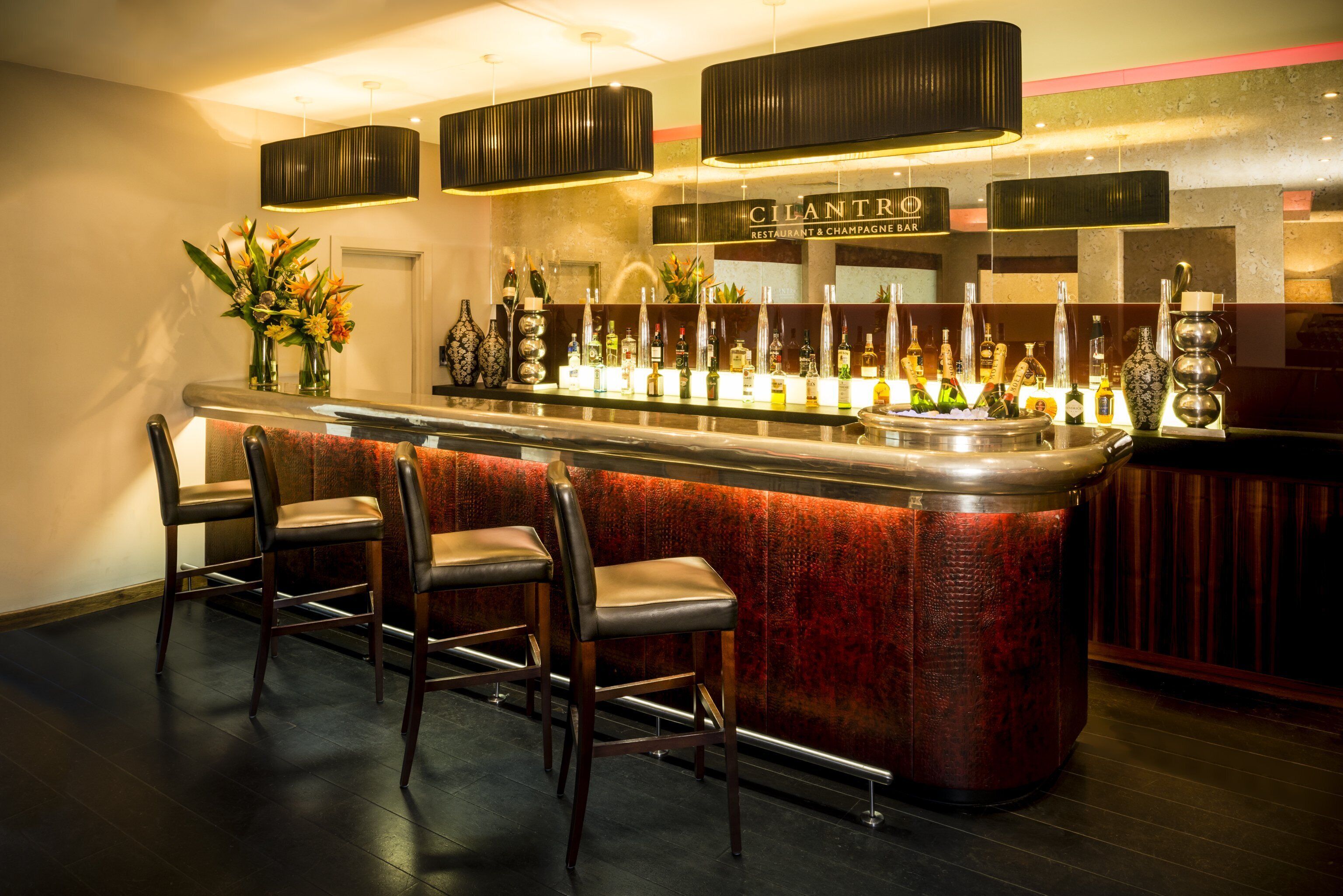 Eine Bar mit einem roten, strukturierten Tresen, drei Barhocker und einer Auswahl an Flaschen und Gläsern.