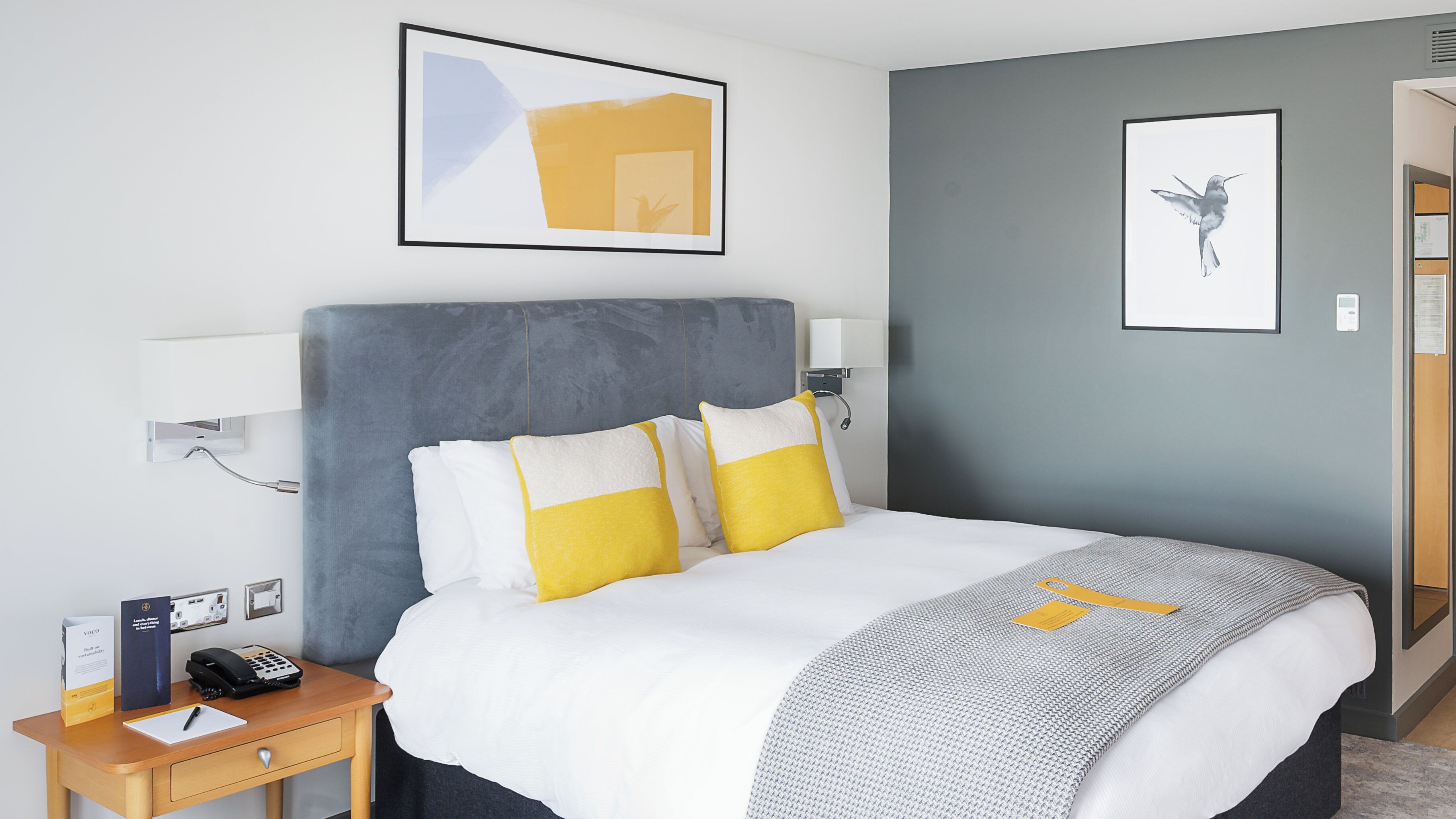 Ein Hotelzimmer mit einem großen Bett, Nachttischen, einem Schreibtisch und einem gerahmten abstrakten Gemälde über dem Bett.