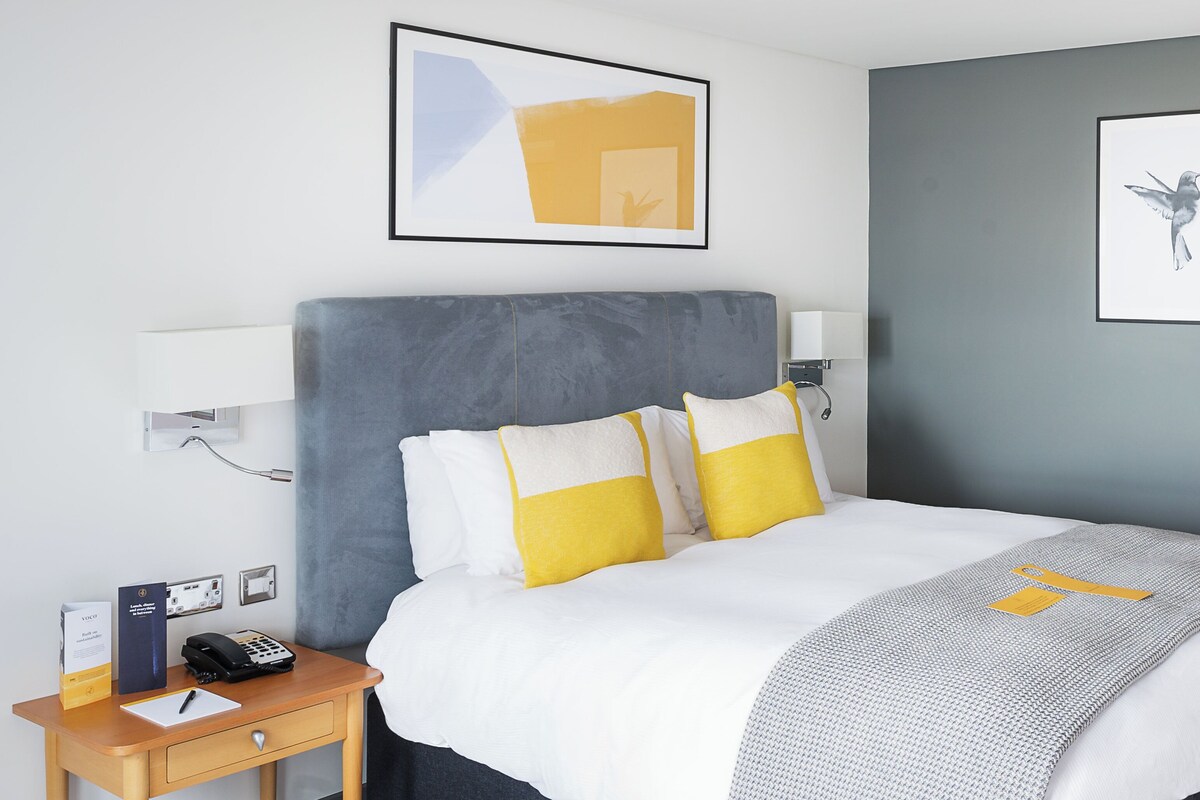 Una habitación de hotel moderna con una cama grande, mesitas de noche, un escritorio y obras de arte en las paredes.