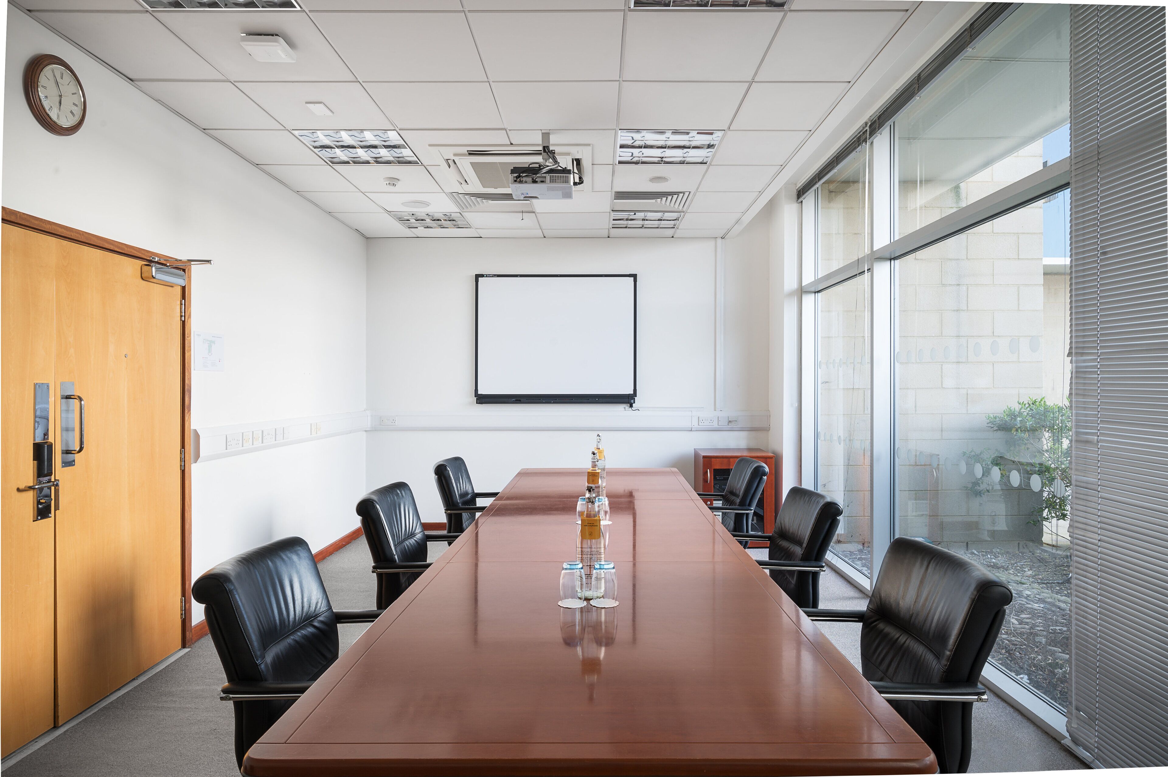 Un salón de conferencias con una mesa larga, seis sillas, una pantalla de proyector y un reloj en la pared.