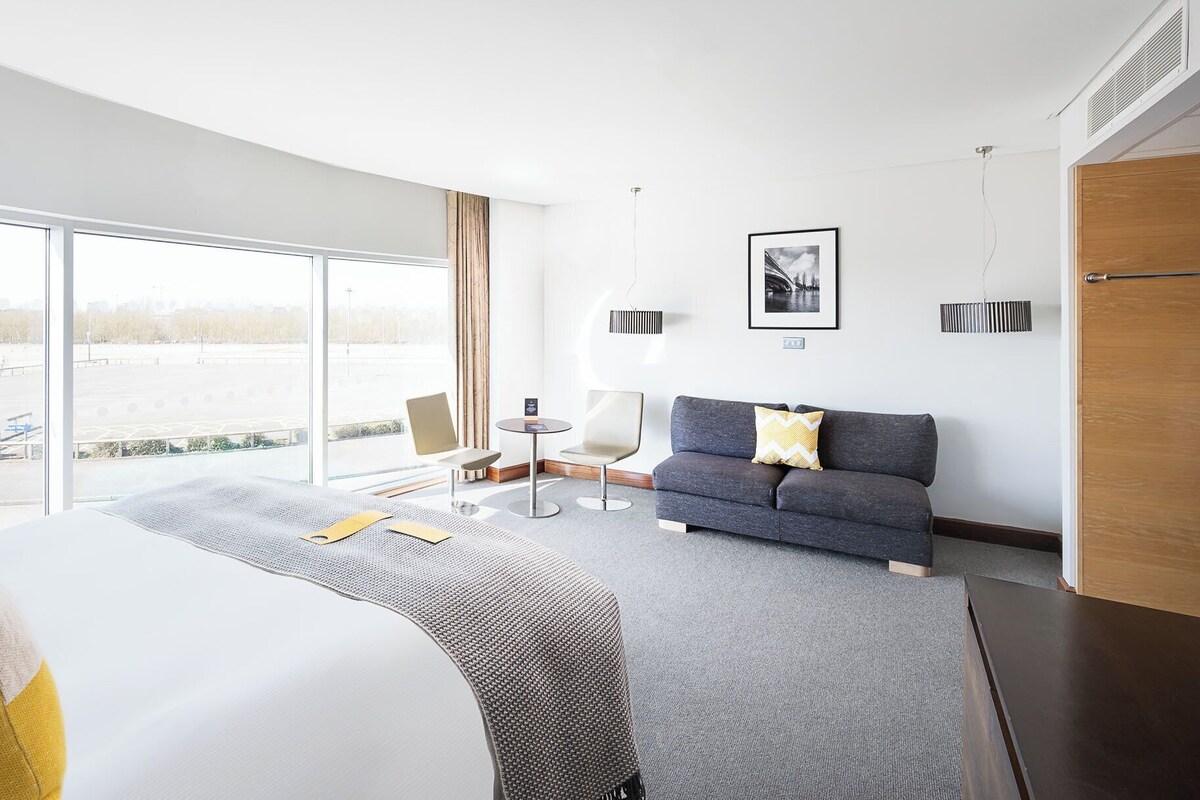 Una habitación de hotel moderna con una cama grande, un sofá gris, una mesita redonda y vistas a un estacionamiento a través de amplios ventanales.