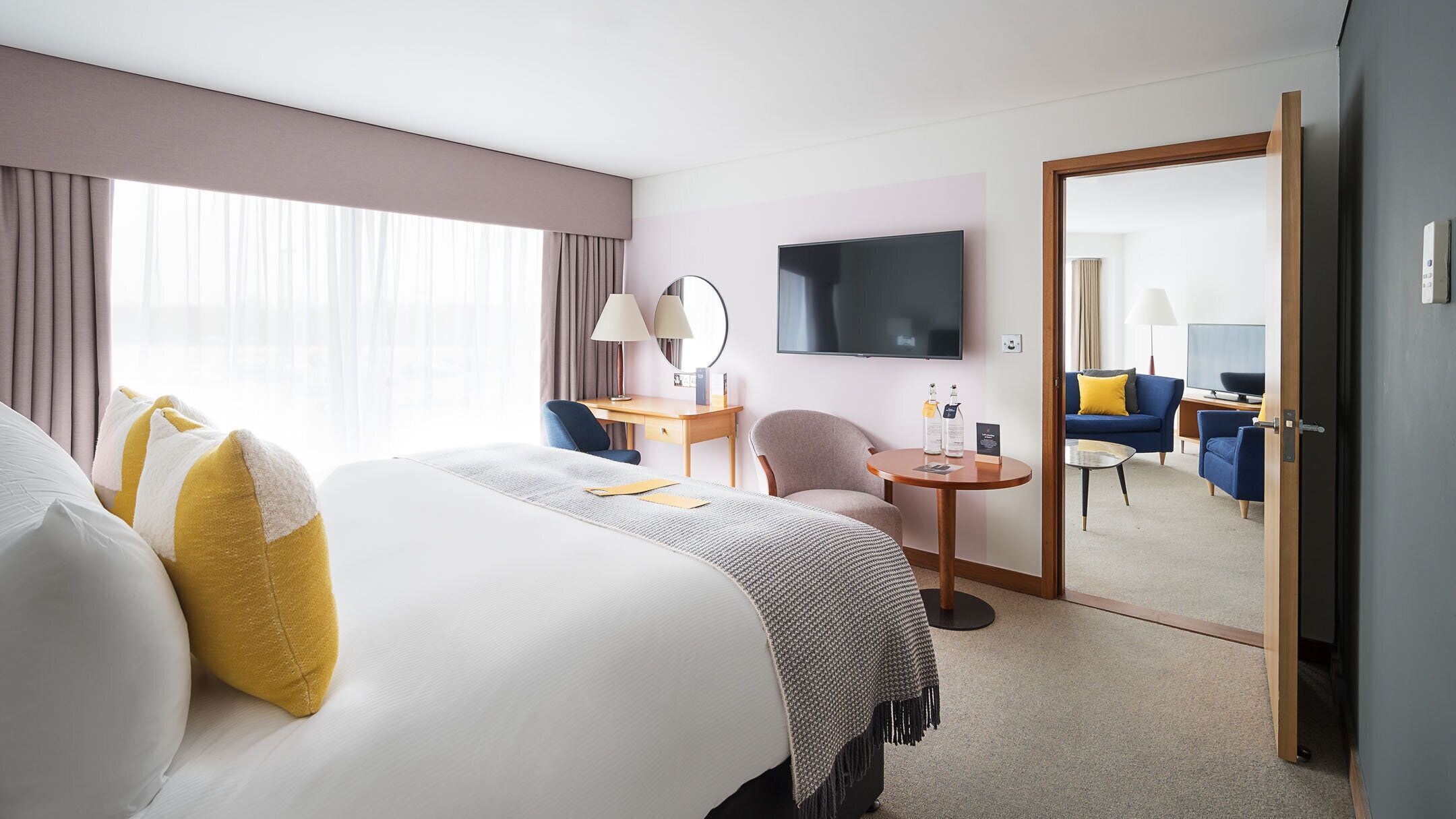 A hotel room with a large bed, a desk, a chair, a small table, a TV, and a mirror.