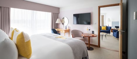 A hotel room with a large bed, a desk, a chair, a small table, a TV, and a mirror.