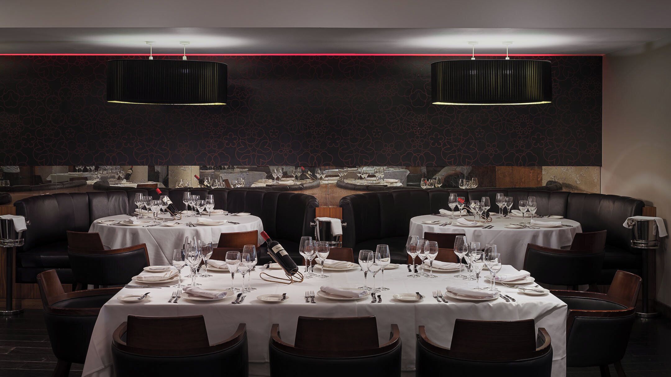 Un restaurante con mesas redondas dispuestas para comer, sillas de cuero negro y una pared oscura con un diseño y una línea roja.