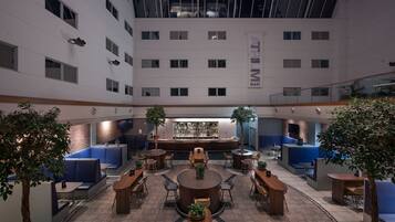 A modern hotel lobby with a central bar, seating areas, and a high ceiling with skylights.