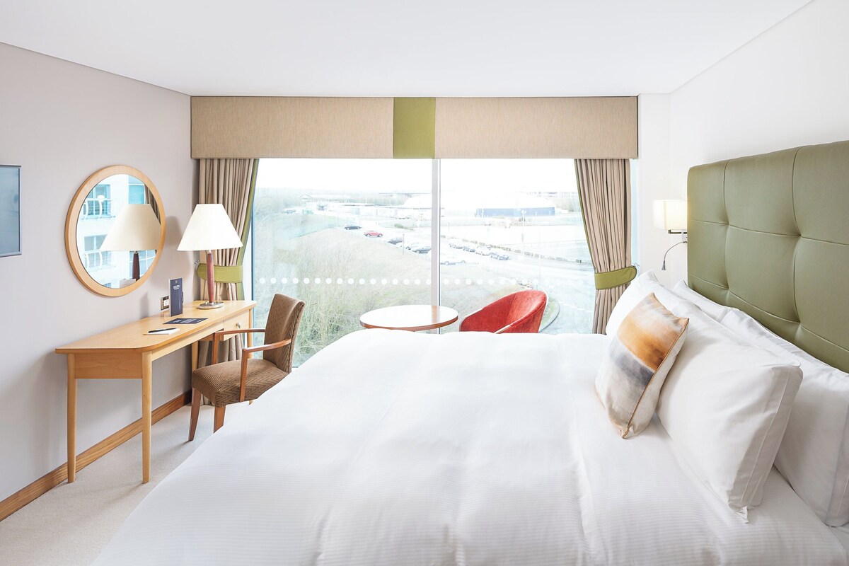 Habitación de hotel con una cama grande, un escritorio, una silla y vista a un estacionamiento a través de la ventana.