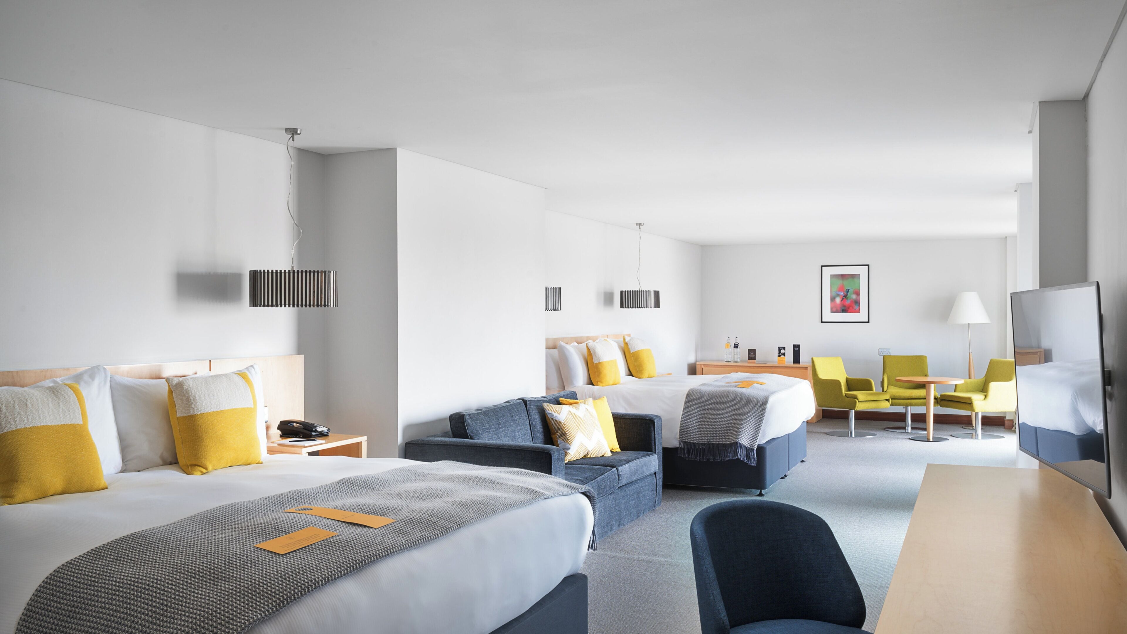 A hotel room with two beds, a sofa, a desk, and a chair. There are bedside tables with lamps and a flat-screen TV.
