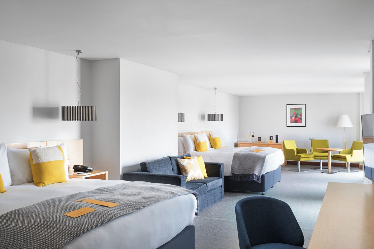 Habitación de hotel con dos camas, un sofá, un escritorio y una silla. Hay dos mesitas de noche con lámparas y un televisor de pantalla plana.