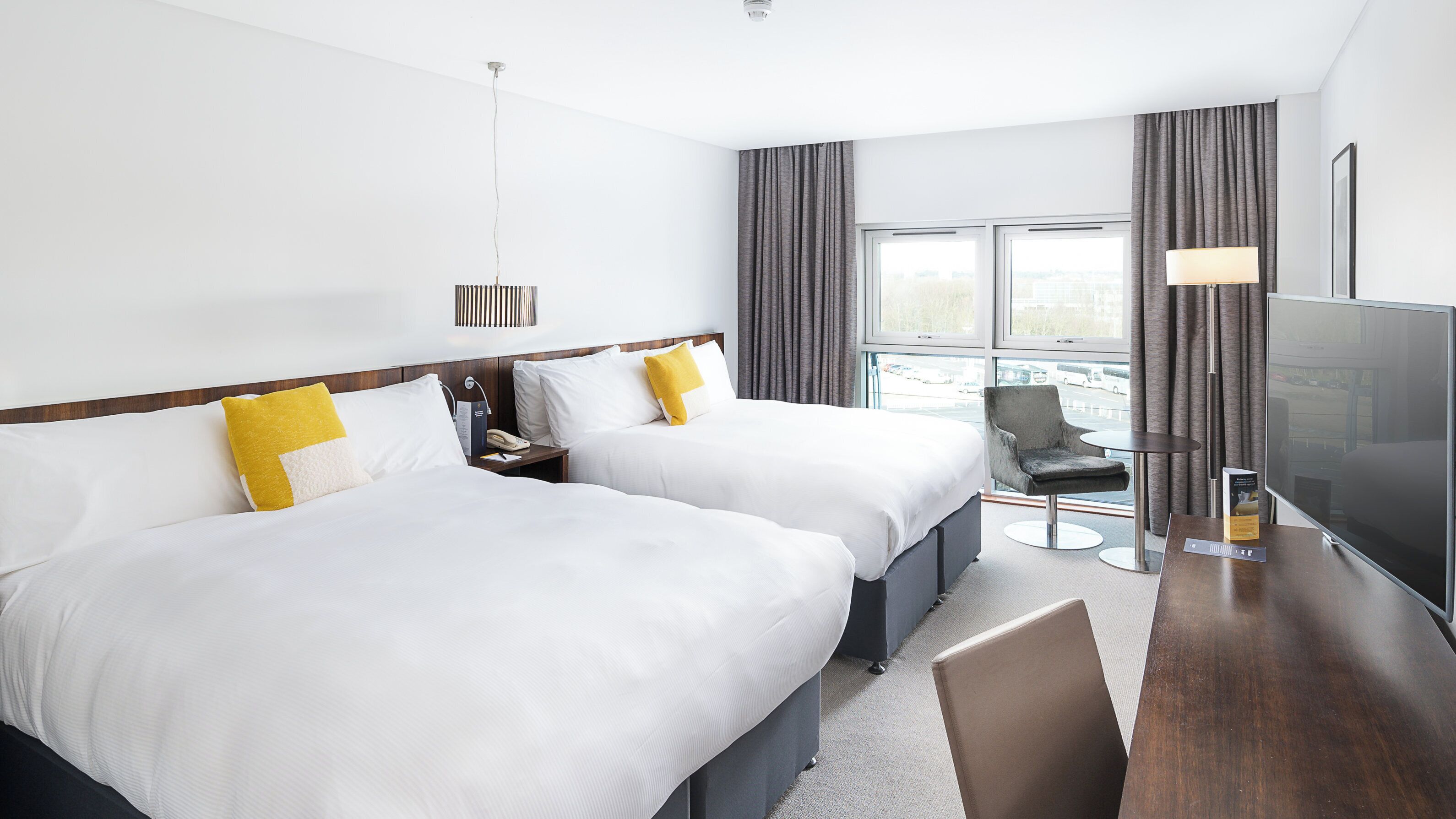 Quarto de hotel com duas camas, uma escrivaninha com cadeira, televisão e janela com cortinas.