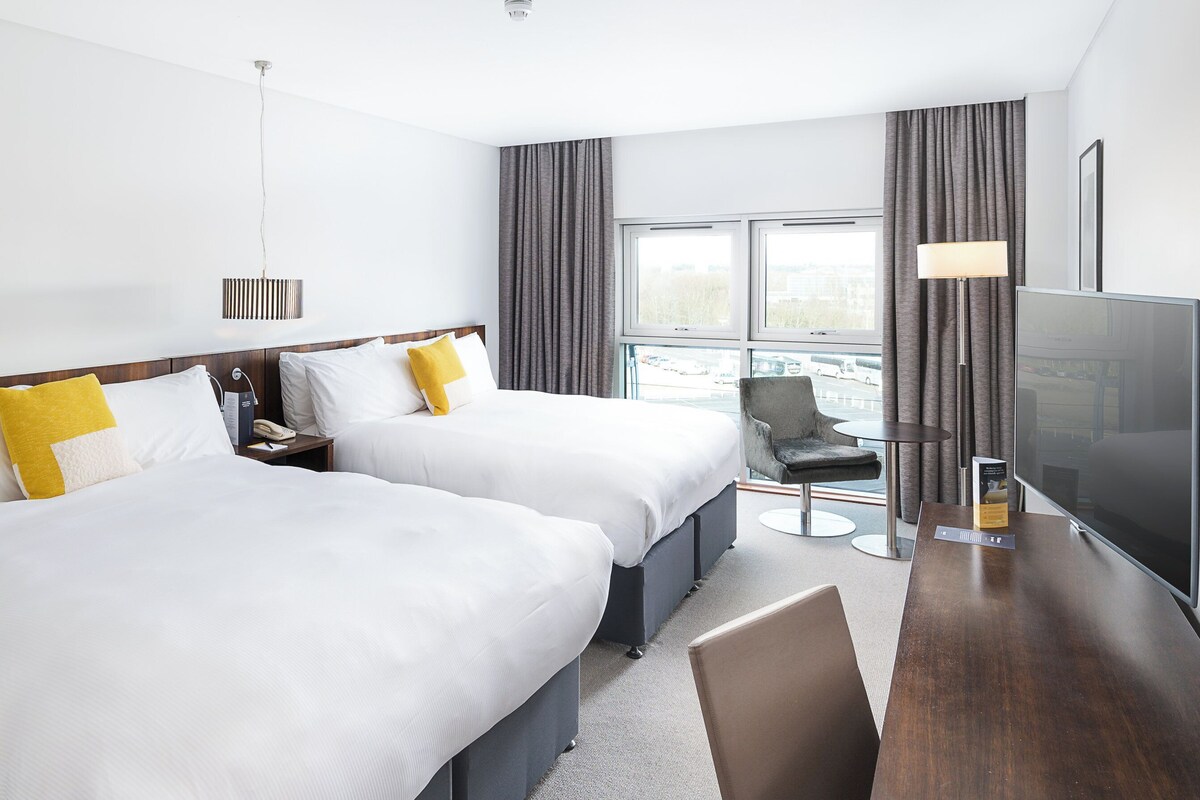 Una habitación de hotel con dos camas, un escritorio con silla, un televisor y una ventana con cortinas.