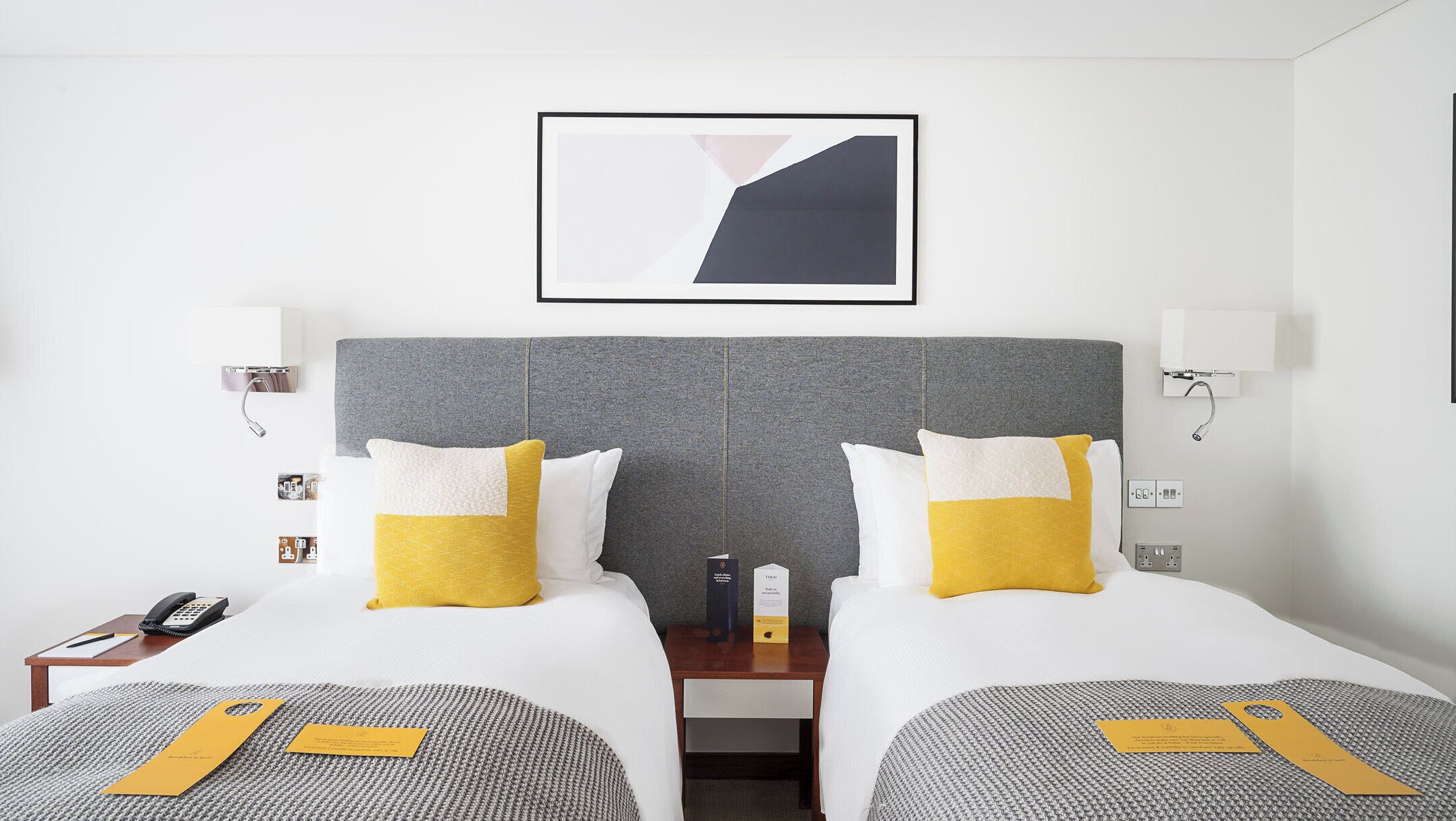 Une chambre d’hôtel avec deux lits, une tête de lit grise, des oreillers blancs et jaunes, une œuvre d’art abstraite encadrée, des tables de chevet avec des lampes et un téléphone sur le côté gauche.