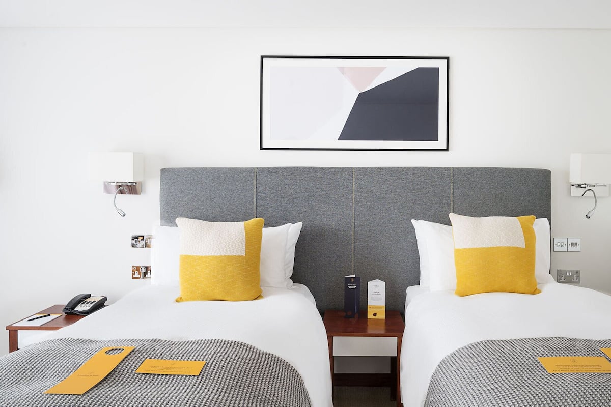 A hotel room with two beds, a grey headboard, white and yellow pillows, a framed abstract artwork, bedside tables with lamps, and a phone on the left side.