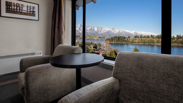 A room with a view of mountains and a lake. There is a round table and two chairs.