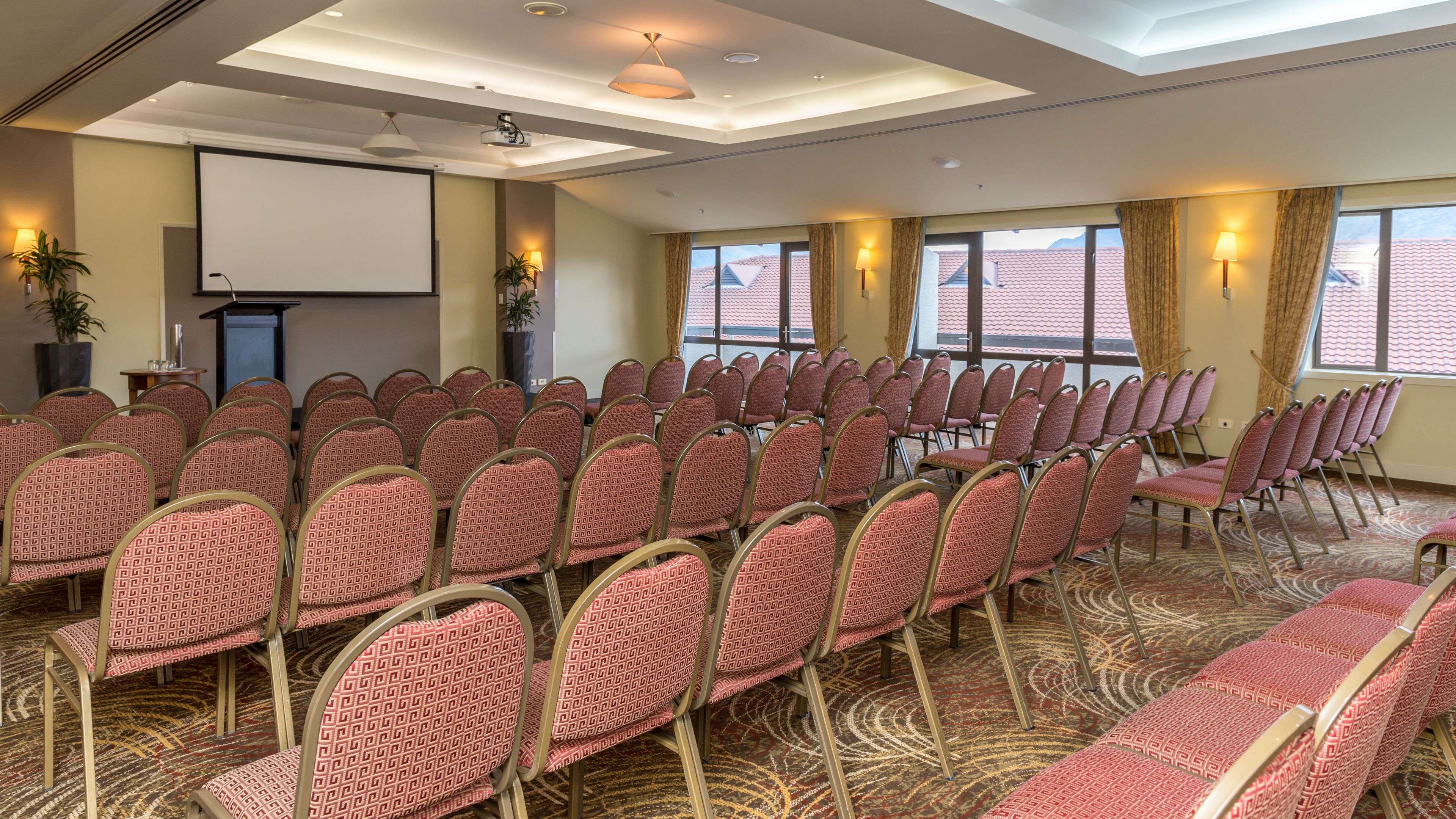 A conference room with rows of chairs, a projector screen, and large windows with curtains.
