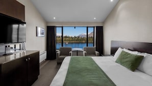 A hotel room with a bed, a desk, a TV, a view of a lake and mountains.