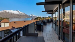 A balcony with a view of a lake and mountains, featuring outdoor seating and a glass railing.