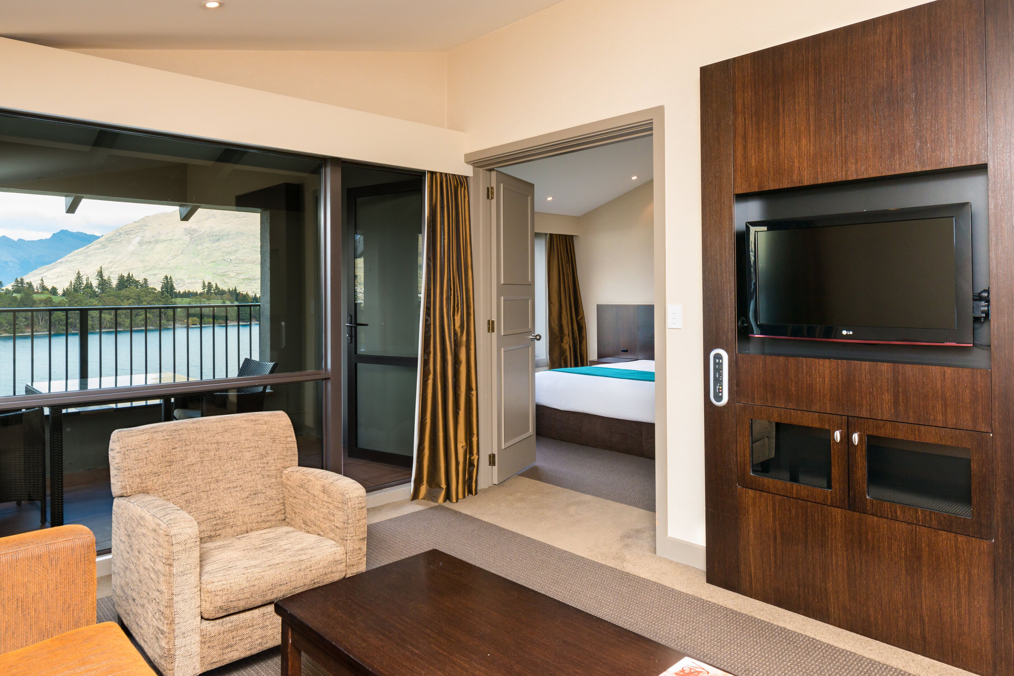 A hotel room with a large TV, a sofa, a bed, and a view of a lake and mountains.