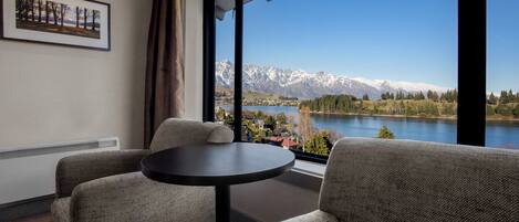A room with a view of mountains and a lake, featuring a round table and two upholstered chairs.