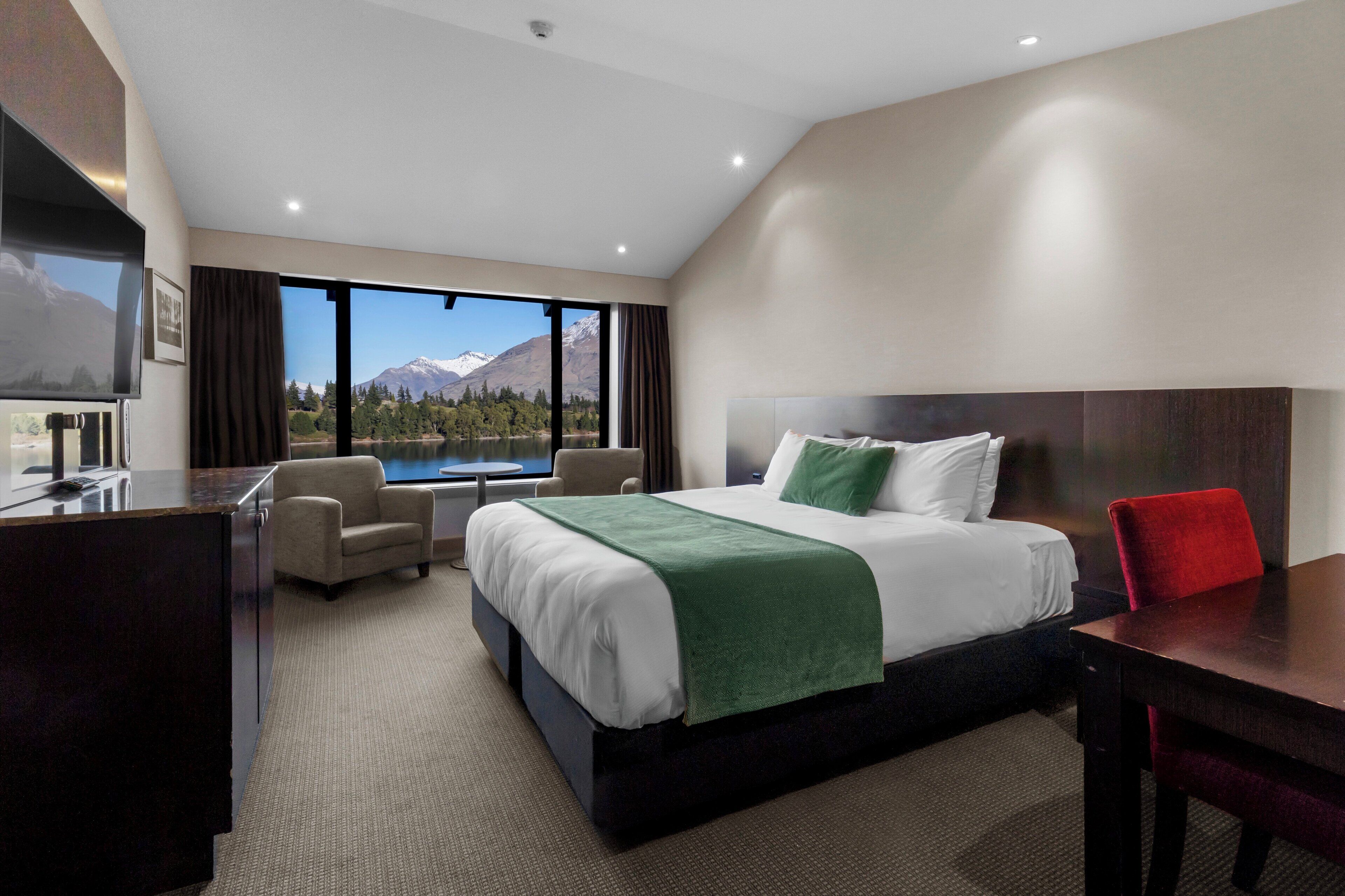 A hotel room with a bed, a desk, a chair, and a view of mountains and a lake.
