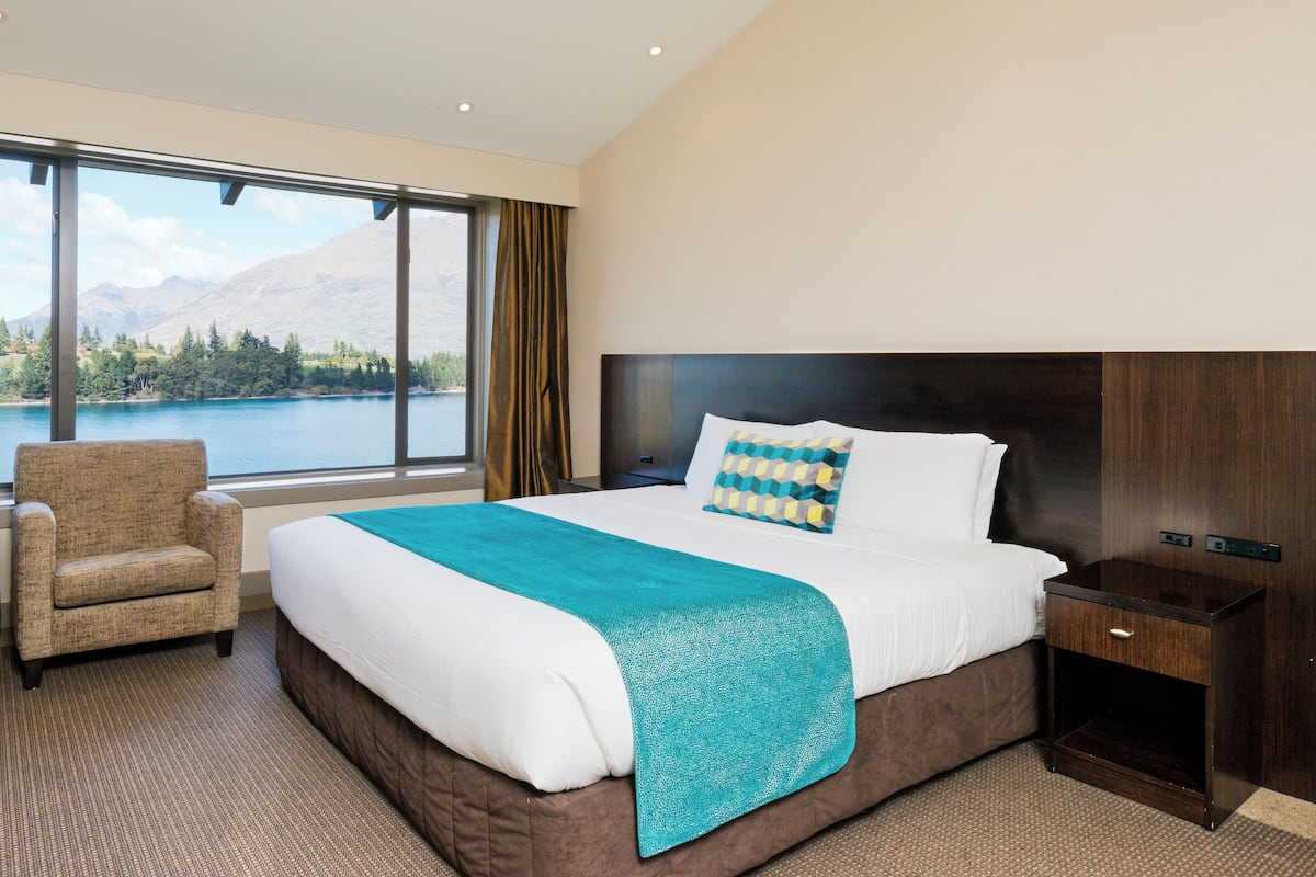 Een hotelkamer met een groot bed, een stoel, een nachtkastje en uitzicht op een meer en bergen.