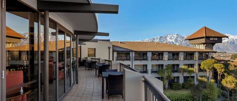 A hotel balcony with outdoor seating, a view of the mountains, and a courtyard with trees.