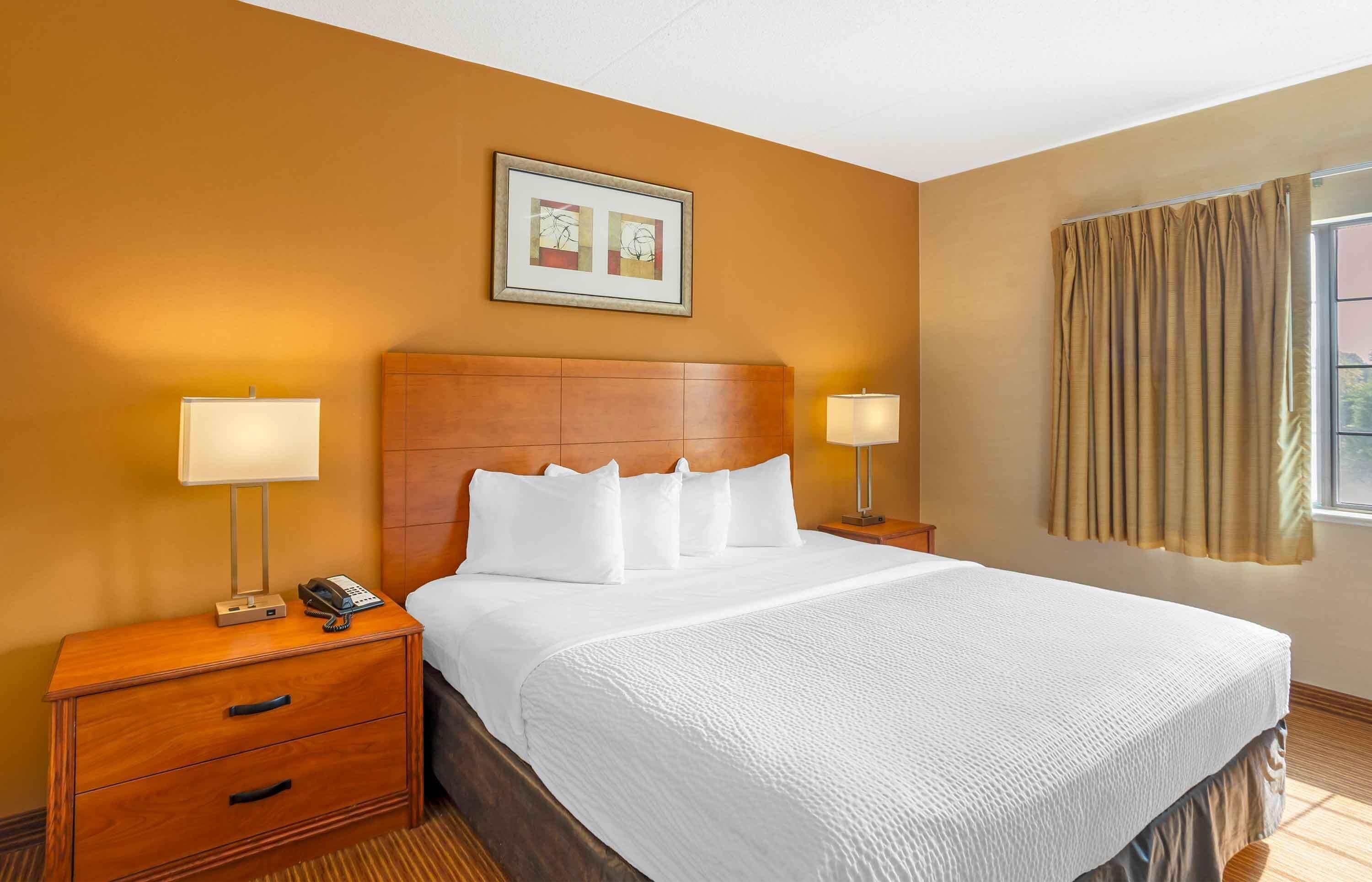 A hotel room with a large bed, wooden headboard, two bedside tables with lamps, a framed picture on the wall, and a window with curtains.