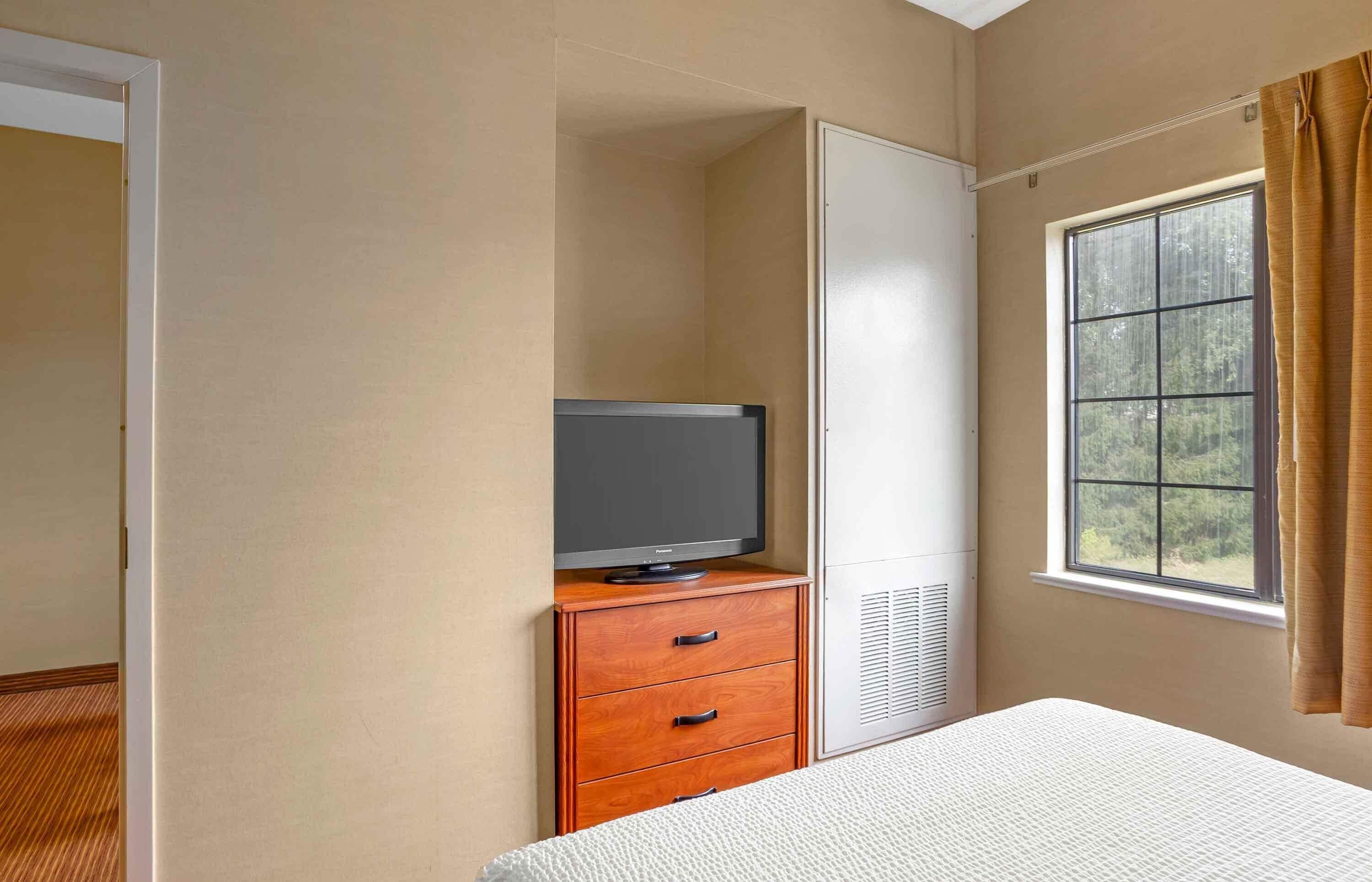 A room with a bed, a television on a wooden dresser, a wardrobe, and a window with curtains.
