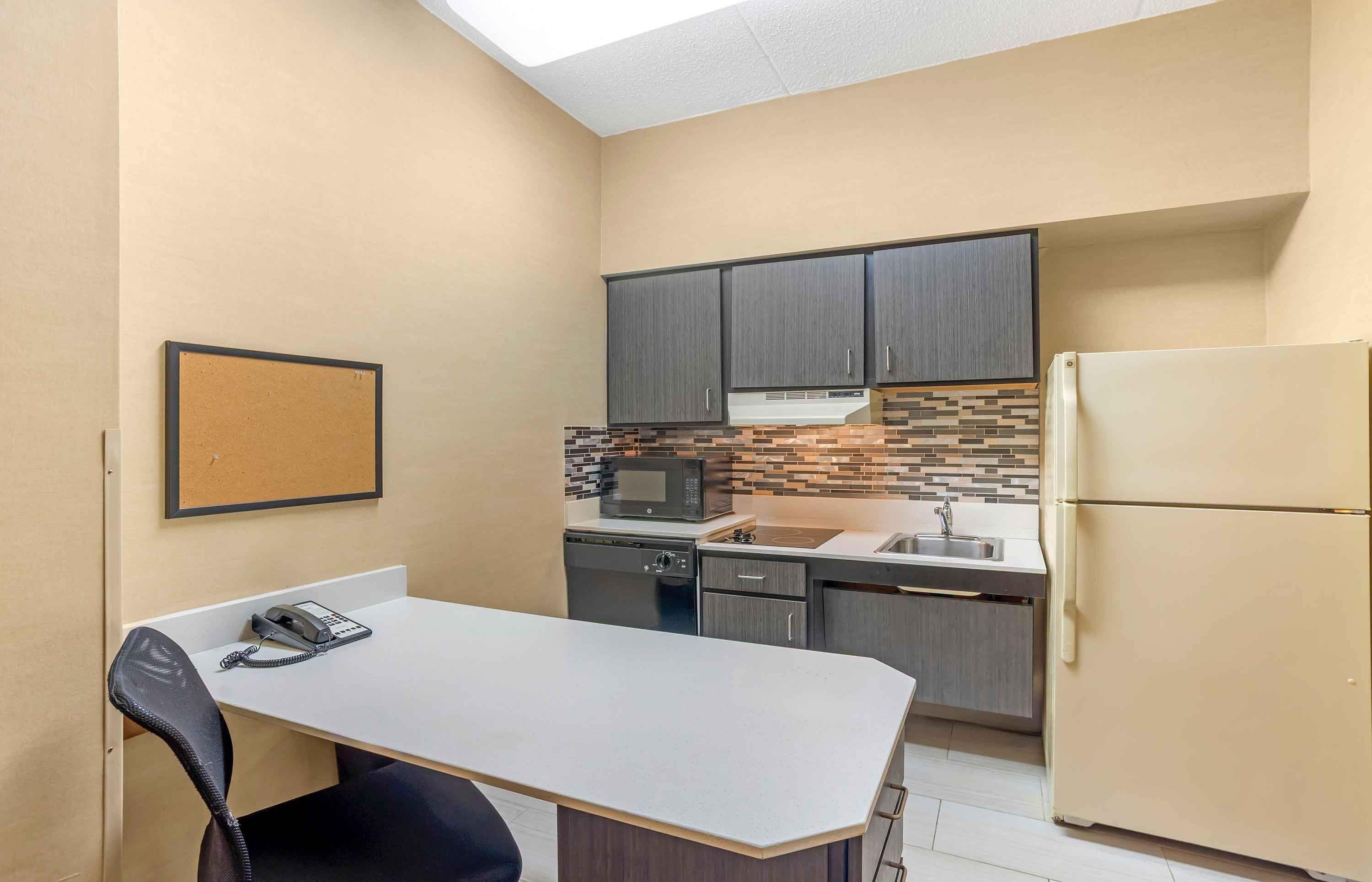 A compact kitchen with a microwave, stove, sink, and refrigerator. There is a small dining table with a chair and a corkboard on the wall.