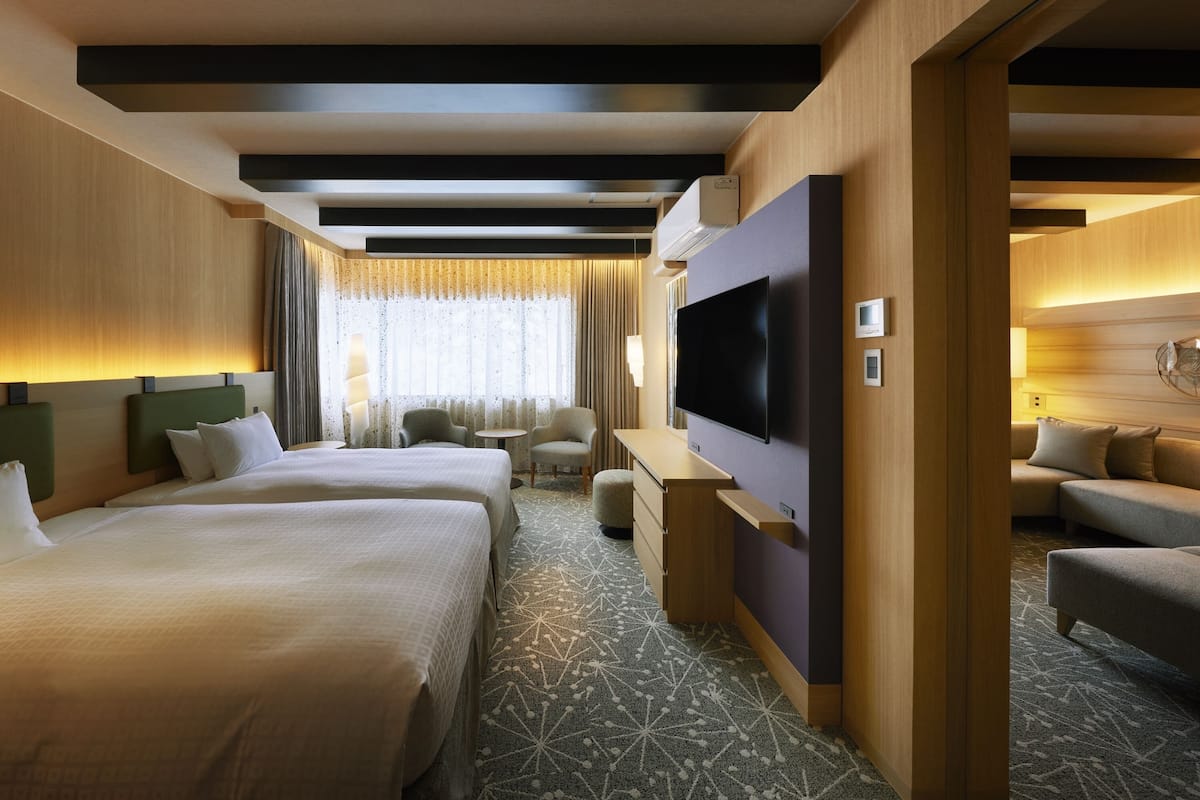 A hotel room with two beds, a desk, a TV, and a seating area.