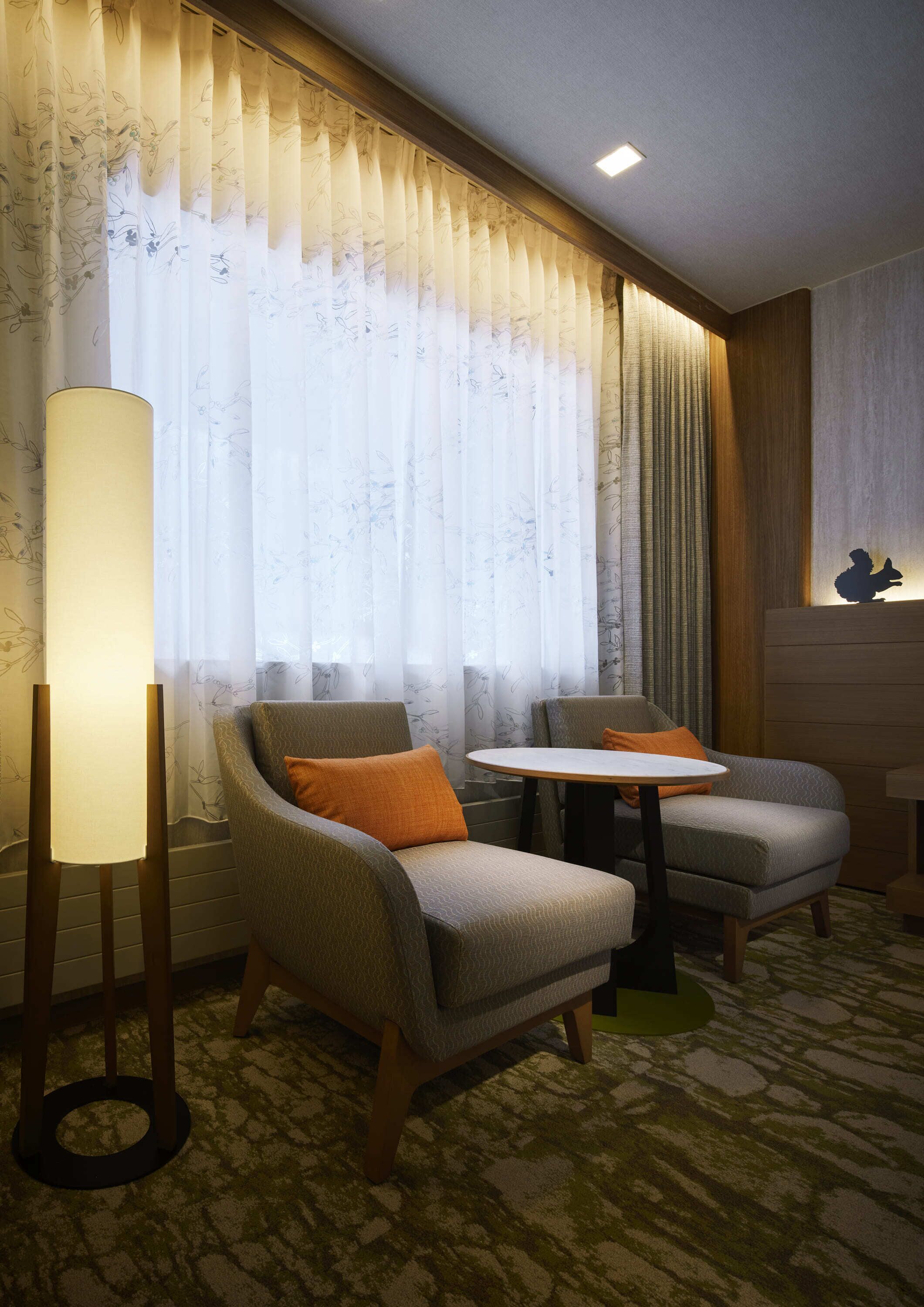 Habitación de hotel con dos sillones, una mesita, una lámpara de pie y un gran ventanal con cortinas.