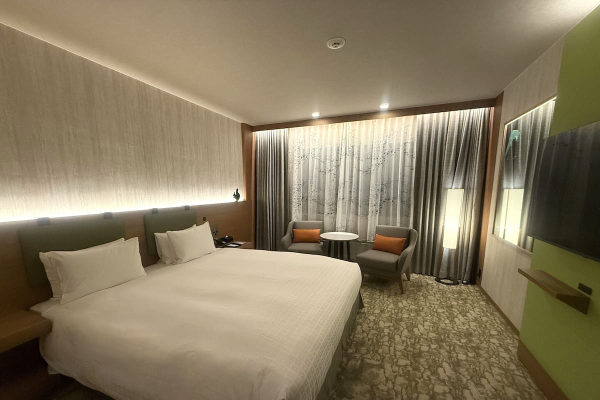 A hotel room with a large bed, a sofa, a small table, and a chair. There is a window with curtains and a view of the outside.