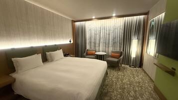A hotel room with a large bed, a sofa, a small table, and a chair. There is a window with curtains and a view of the outside.