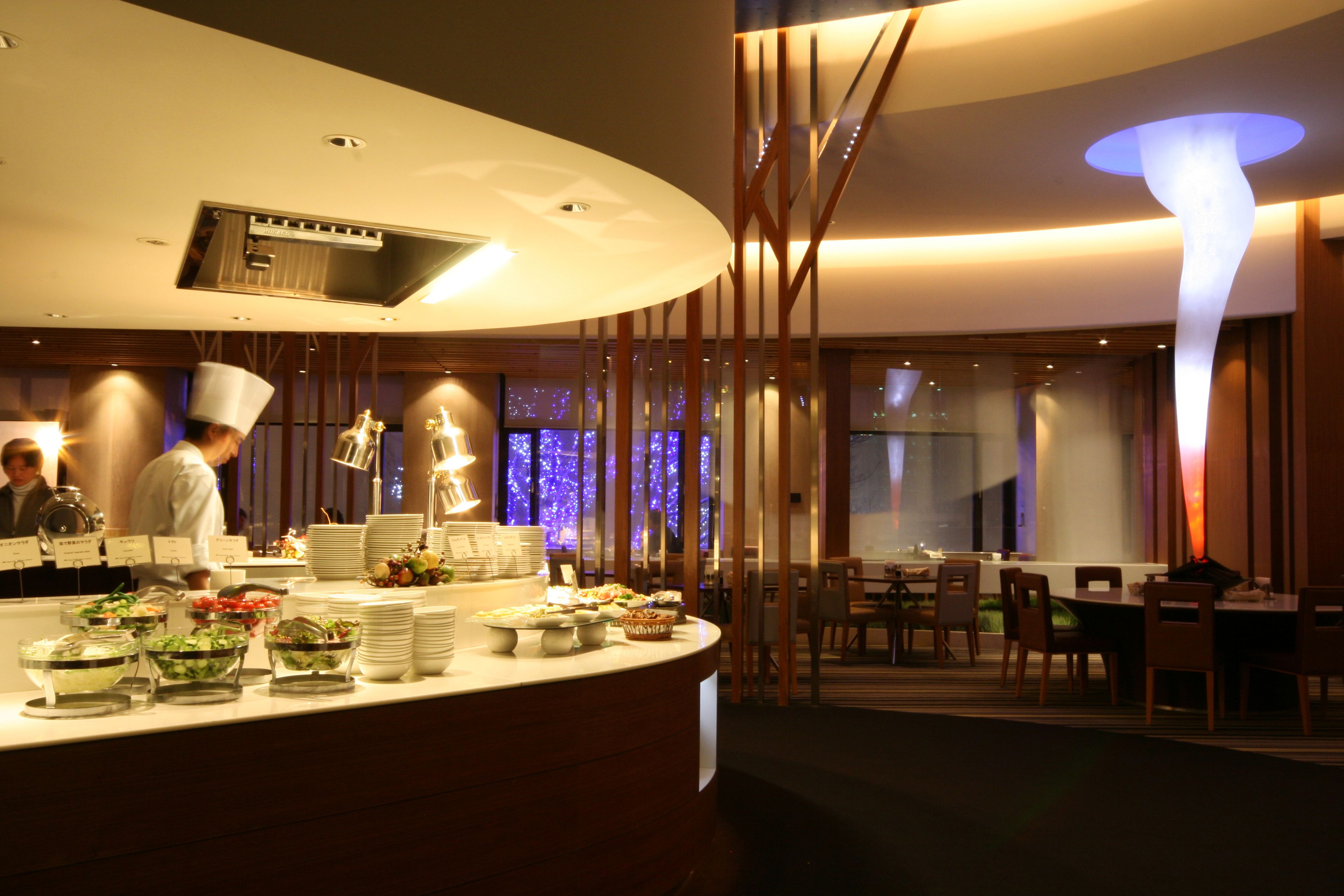 A modern restaurant with a curved counter displaying various dishes, a chef in uniform, and a contemporary interior design.