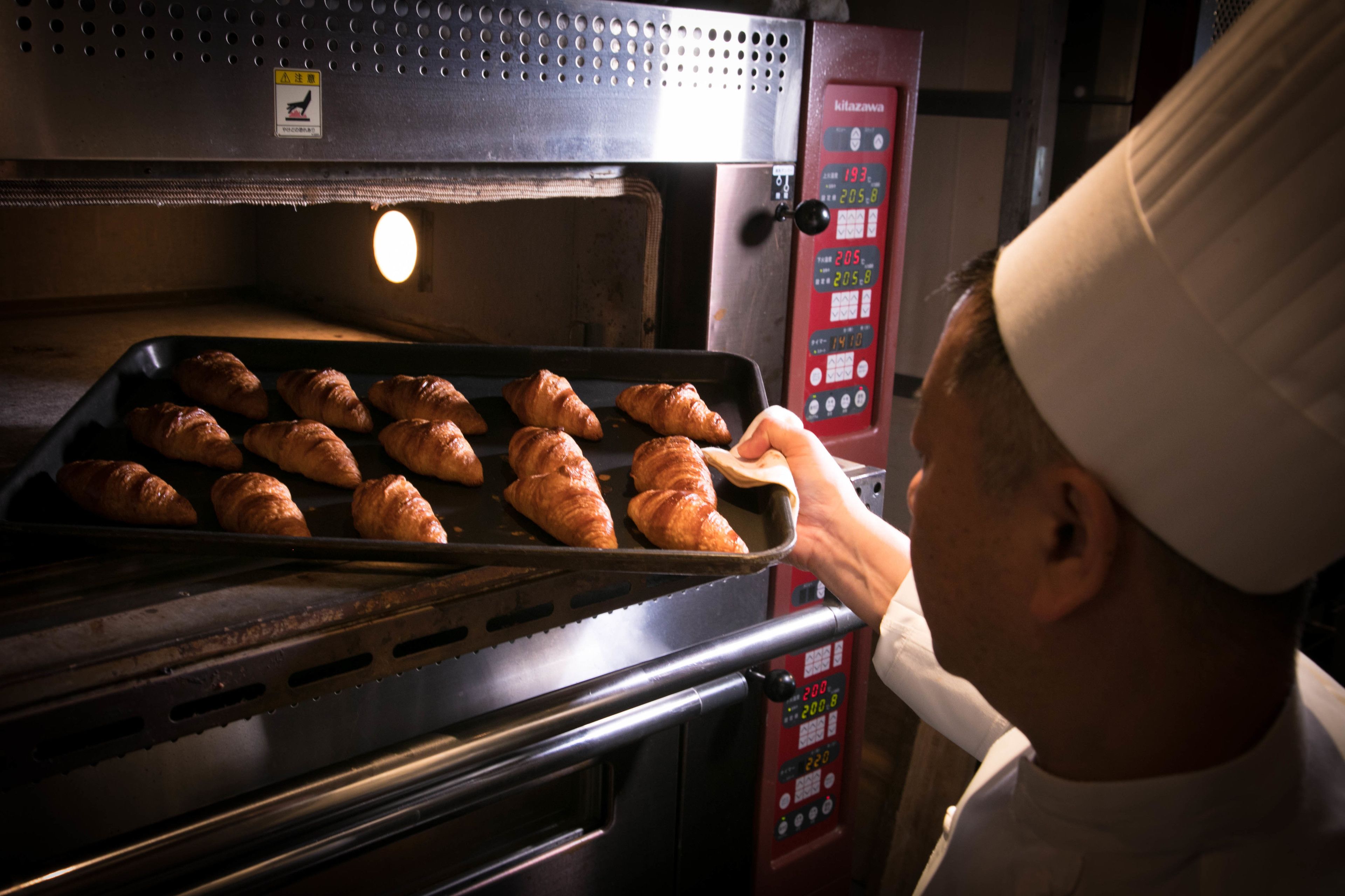 Eine Person mit Kochmütze legt ein Tablett mit Croissants in einen Industriebeis.