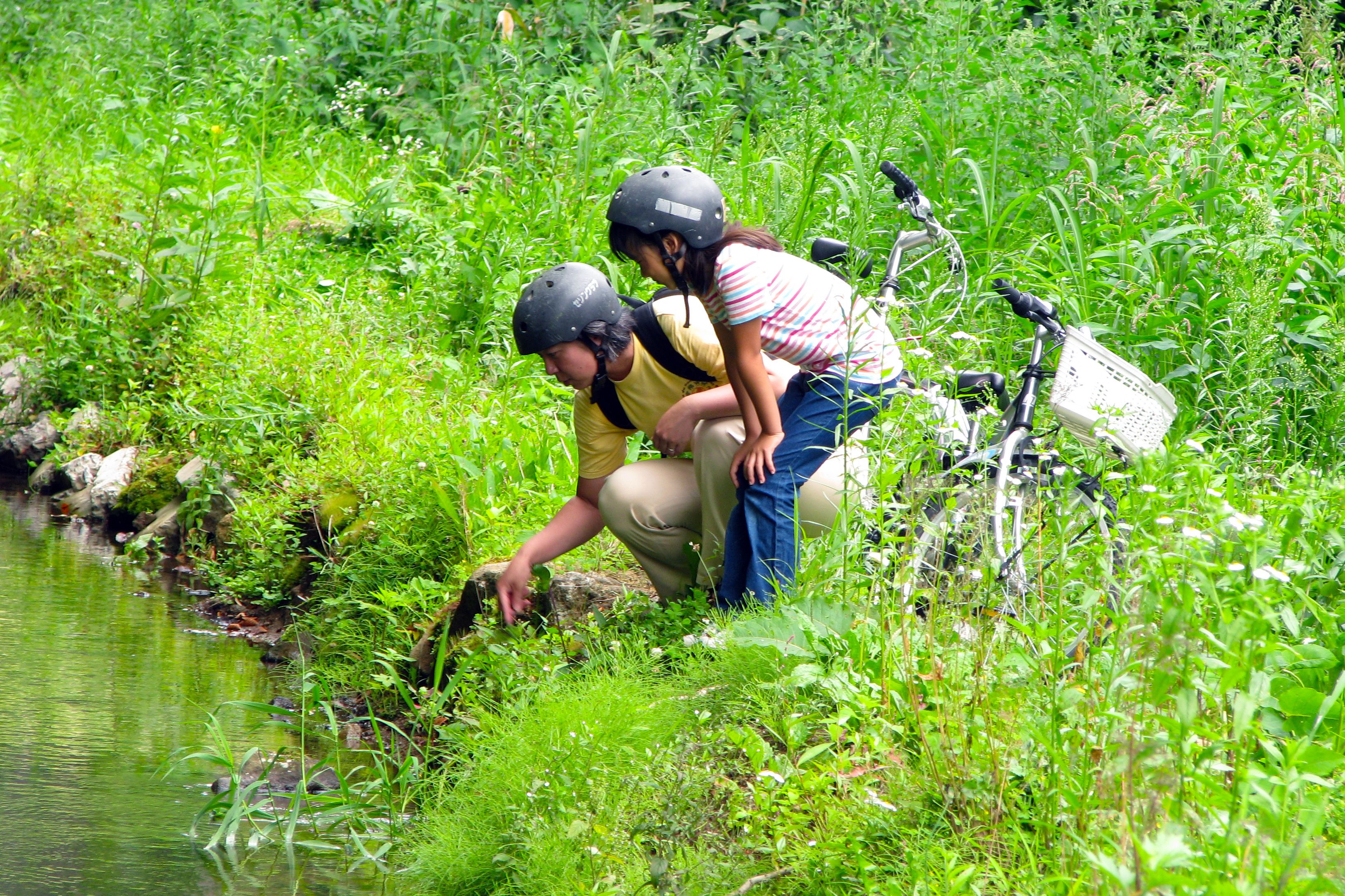 Dos personas con casco cerca de un cuerpo de agua rodeado de vegetación.