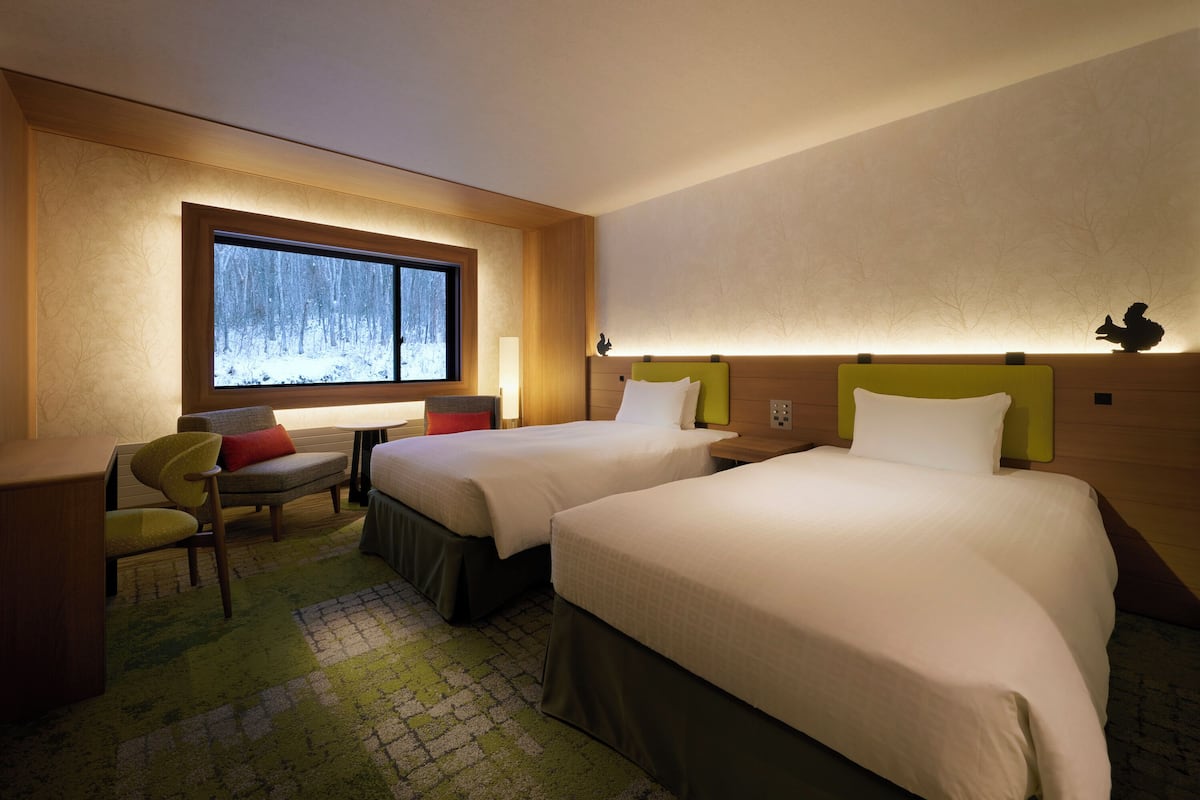 A hotel room with two beds, a desk, a chair, a window with a view of a snowy forest, and a lamp.